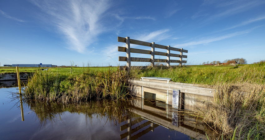 Foto van weiland, met vooraan een hek en een sloot en in de achtergrond weg en bebouwing